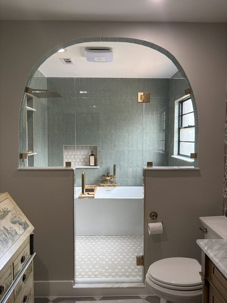 After: arched glass wet room with sage green vertical tile, freestanding soaking tub, and brass rain shower