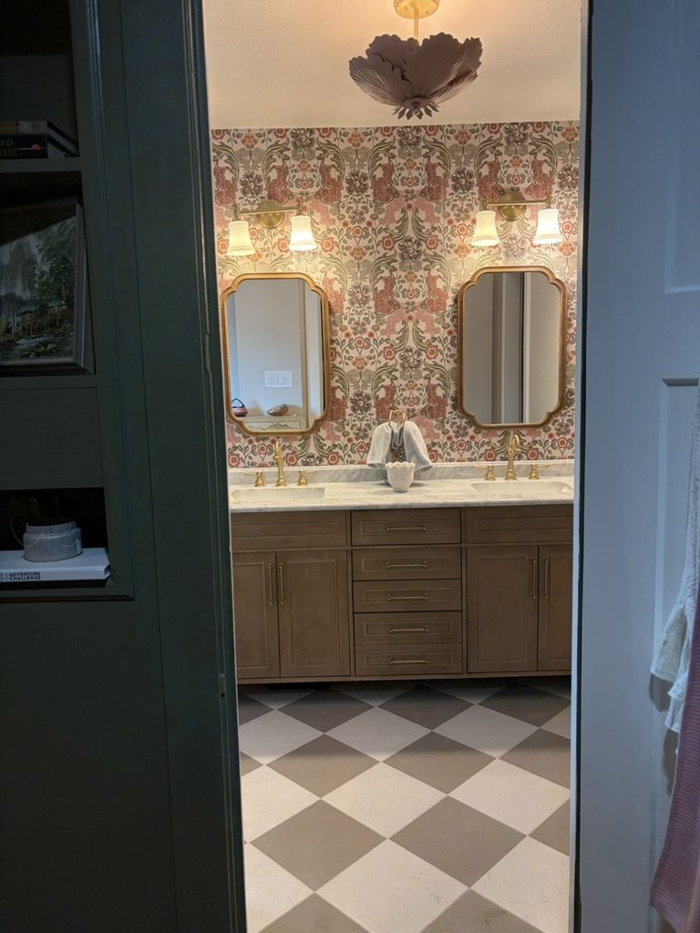 After: view from a green-paneled bedroom into a remodeled bathroom with a double vanity, botanical wallpaper, and checkerboard floor