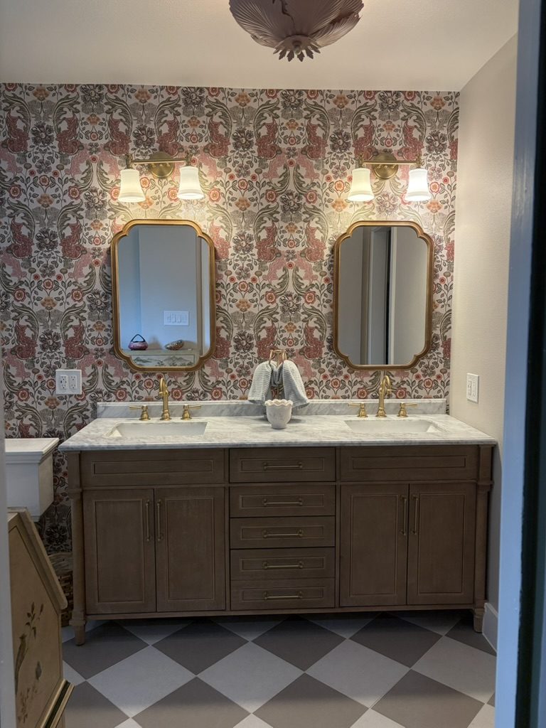 After: wood double vanity with marble top on an oversized checkerboard tile floor, set against blush and sage botanical wallpaper